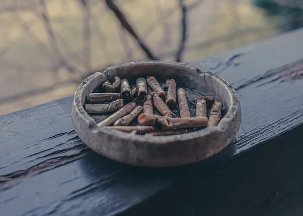 A clay ashtray with butts on a wooden terrace beam. Smoking on the street. Remnants of cigarettes. Nicotine addiction. Ashtray with cigarette butts. Deadly dangerous habit.