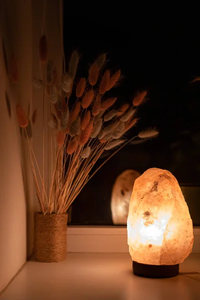 Warm light from a salt lamp illuminating a bouquet of lagurus in the evening. Warm light. Room lighting. Home comfort. The benefits of a salt lamp. Dry flowers in the interior. Apartment decoration.