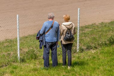 Yaşlı bir adam ve kadın sırtı kameraya dönük bir şekilde çitin önünde durup ileriye bakarlar. Evli çift dikiz aynası. Sırtını kameraya dön. Emeklilikte boş zaman. Birlikte yürüyün. Yaşlı bir çift. Emeklilerin hayatı. Yaşlı ebeveynler.