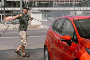 self-servis araba yıkama. Şoför arabasını yıkar. Erkek işi. Şapkalı bir adam kırmızı bir hatchback 'i basınçlı suyla yıkıyor. Arabaya dikkat et..