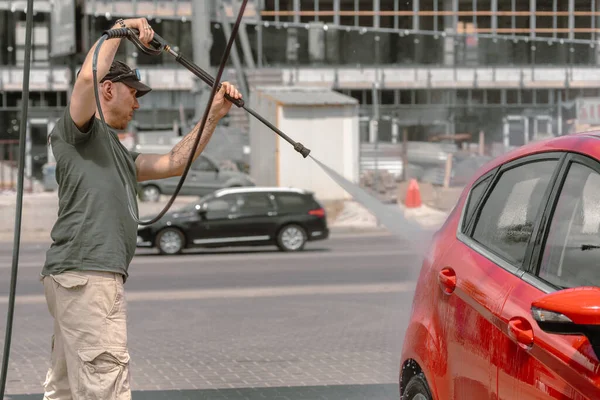 self-servis araba yıkama. Şoför arabasını yıkar. Erkek işi. Şapkalı bir adam kırmızı bir hatchback 'i basınçlı suyla yıkıyor. Arabaya dikkat et..