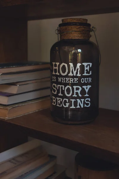 A glass jar with illumination and an inscription about home on a bookshelf. The original lamp. Glass jar with illumination. Inscription about home. Bookshelf with home decor. A cozy corner. White letters.