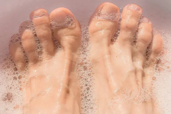 Ev spa tedavisi sırasında köpüklü kadın ayakları. Yorgun ayaklar için banyo. Köpük kokusundan zevk alın. Su işlemleri sırasında iyi eğlenceler. Kişisel bakım. Sağlıklı bir yaşam tarzı. Islak ayaklar. Saydam tırnak cilası.
