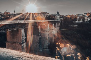 Köprünün kenarından eski Ukrayna şehri Kamianets-Podilskyi 'ye gün batımı. Kontrastlı güneş ışığı altında bir şehir. Avrupa 'nın mimarisi. Ukrayna 'da turizm. Parapet 'in sütunlarındaki güneş ışınları. Geometri ve şehir mimarisinin perspektifi.