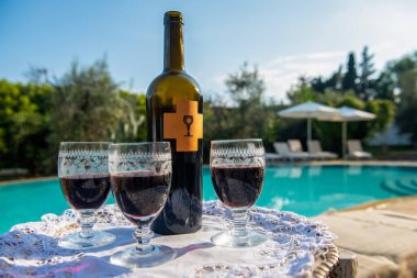 Bir şişe mükemmel kırmızı şarap ve çok güzel dekore edilmiş üç bardak yüzme havuzunun arka planında bir tepside duruyor.