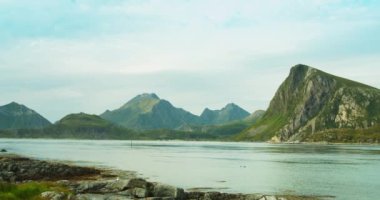 Güneşli bir günde göl ve dağlar manzaralı. Ön planda kayalar ve çimenler var. Lofoten Adaları, Norveç fiyortlarının güzel doğası..