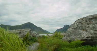 Göl ve dağların manzarası bulutlu bir günde manzaralı. Ön planda kayalar ve çimenler var. Lofoten Adaları, Norveç fiyortlarının güzel doğası..