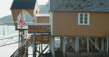 Güneşli bir günde Norveç bayrağı rüzgarda dalgalanıyor. Marinadaki geleneksel sahil kulübesi. Reine, Lofoten Adaları