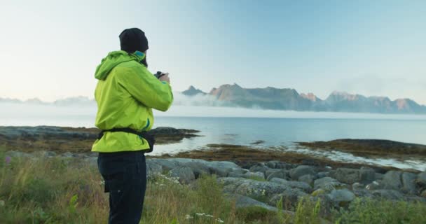 一位穿着鲜艳绿色夹克的摄影师拍摄了美丽的挪威峡湾的照片 那个人拿着摄像机站在湖边 晴天背景中的低云 — 图库视频影像