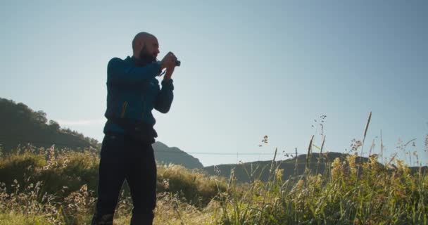 一位穿着蓝色运动衫的摄影师拍摄了美丽的挪威峡湾的照片 那个男人站在高高的草地上 手里拿着相机 在背景中 多山的地形在阳光灿烂的早晨 太阳耀斑出现 慢动作视频 — 图库视频影像