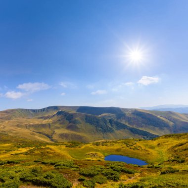 Parlak güneşli bulutlu bir gökyüzünün altında dağda küçük mavi bir göl.