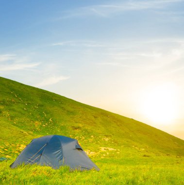 Güneş batarken yeşil dağ yamacına yakın turistik çadır, yaz tatili manzarası.