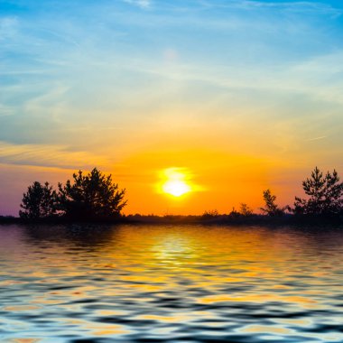 Sakin bir gölün üzerinde dramatik bir gün batımı ve sahilde orman silueti.