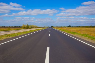Mavi bulutlu gökyüzünün altında, yeşil ovaların arasında uzun asfalt yol
