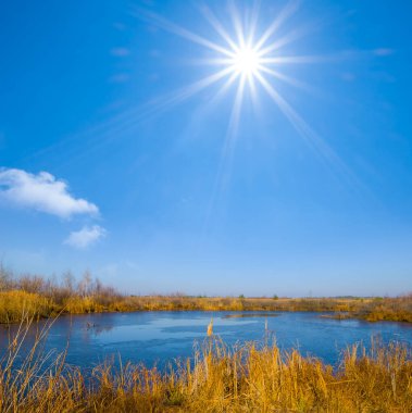 Aydınlık güneşli bir günde, kıyıda kuru kamışlı ovaların arasında küçük bir göl.