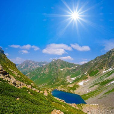 Parlak güneşin altında dağ zincirinin arasında küçük sakin bir göl.