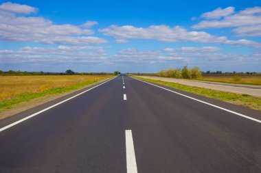 Mavi bulutlu gökyüzünün altında, yeşil ovaların arasında uzun asfalt yol