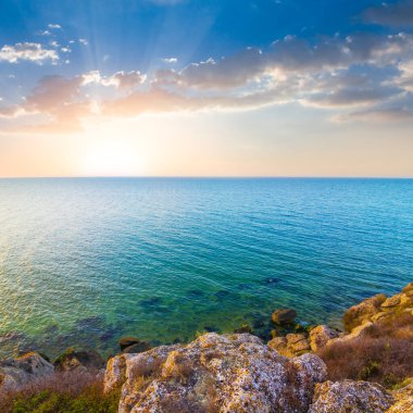Gün batımında mavi bulutlu gökyüzünün altında deniz koyunun taşlı kıyıları.