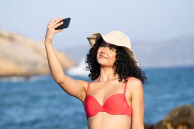 Sahilde genç ve güzel bir kız. Plaj şapkalı. Telefon kamerasıyla selfie çekiyor.