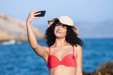 Sahilde genç ve güzel bir kız. Plaj şapkalı. Telefon kamerasıyla selfie çekiyor.