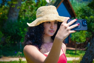 Sahilde genç ve güzel bir kız. Plaj şapkalı. Telefon kamerasıyla selfie çekiyor.