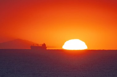 Gün batımında ve gün doğumunda ufukta kargo gemilerinin siluetleri beliriyor.