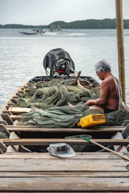 Chantaburi, Tayland, 21 Mart 2023: Tahta bir kayıktaki bir balıkçı, uzun kuyruklu bir tekne uzaklardan geçerken ağlarını onarır..