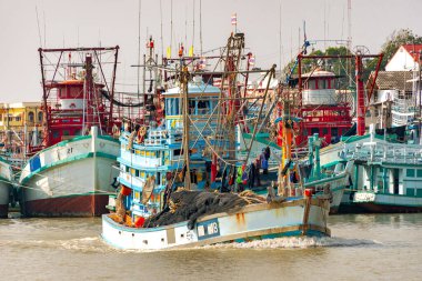 Yoğun bir balıkçı limanında, çeşitli boylarda ve renklerde çok sayıda balıkçı teknesi bir su yoluna demir atar. Balıkçılık endüstrisiyle ilişkili tipik donanım ve ağlarla donatılmışlardır..
