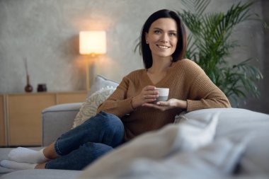 Mutlu, orta yaşlı bir kadın oturma odasında rahat bir koltukta, elinde bir fincan siyah çay ya da kahveyle oturuyor. Huzurlu 30 'lu yaşlardaki kadın stressiz, sakin, evde tek başına eğleniyor.