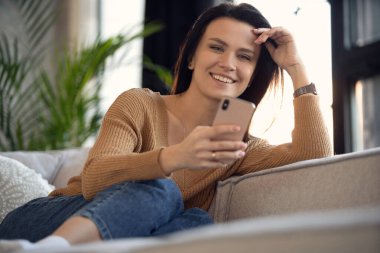 Milenyum boyunca rahat koltukta rahat rahat uzanan akıllı telefonu elinde tutan mutlu bir kadın. Gülümseyen 30 'lu yaşlardaki kadınlar sosyal ağlarda sohbet ediyorlar, komik videolar izliyorlar, evde mobil uygulamalar kullanıyorlar.