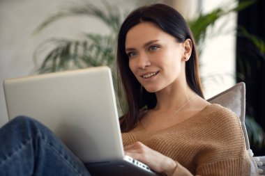 Dizüstü bilgisayar kullanan gülümseyen bir kadın, evdeki koltukta oturuyor, güzel bir bayan alışveriş yapıyor ya da sosyal ağda sohbet ediyor, eğleniyor, film izliyor, bilgisayar projesinde serbest çalışıyor.