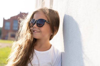 Moda çocuğu konsepti. Beyaz duvara karşı parlak kıyafetler ve güneş gözlüğü takan şık bir kız çocuğu.