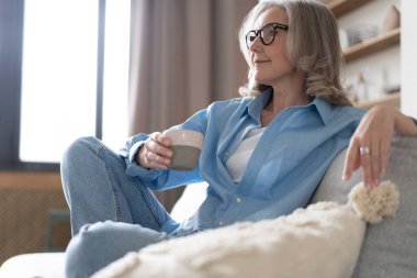 Mutlu, güzel, rahat, olgun, gri saçlı kadın evdeki kanepede dinlenirken kahve içiyor. Gülümseyen orta yaşlı bayan modern oturma odasındaki kanepede dinlenmekten zevk alıyor..
