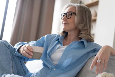 Mutlu, güzel, rahat, olgun, gri saçlı kadın evdeki kanepede dinlenirken kahve içiyor. Gülümseyen orta yaşlı bayan modern oturma odasındaki kanepede dinlenmekten zevk alıyor..