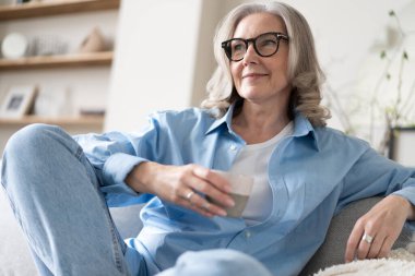 Mutlu, güzel, rahat, olgun, gri saçlı kadın evdeki kanepede dinlenirken kahve içiyor. Gülümseyen orta yaşlı bayan modern oturma odasındaki kanepede dinlenmekten zevk alıyor..