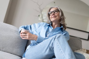 Mutlu, güzel, rahat, olgun, gri saçlı kadın evdeki kanepede dinlenirken kahve içiyor. Gülümseyen orta yaşlı bayan modern oturma odasındaki kanepede dinlenmekten zevk alıyor..