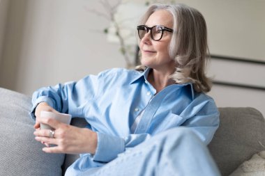Mutlu, güzel, rahat, olgun, gri saçlı kadın evdeki kanepede dinlenirken kahve içiyor. Gülümseyen orta yaşlı bayan modern oturma odasındaki kanepede dinlenmekten zevk alıyor..