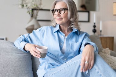 Mutlu, güzel, rahat, olgun, gri saçlı kadın evdeki kanepede dinlenirken kahve içiyor. Gülümseyen orta yaşlı bayan modern oturma odasındaki kanepede dinlenmekten zevk alıyor..