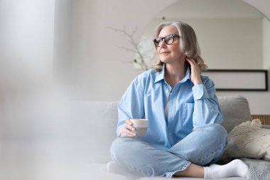 Mutlu, güzel, rahat, olgun, gri saçlı kadın evdeki kanepede dinlenirken kahve içiyor. Gülümseyen orta yaşlı bayan modern oturma odasındaki kanepede dinlenmekten zevk alıyor..