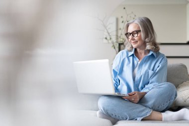 50 'li yaşlardaki gülümseyen olgun bir kadın elinde dizüstü bilgisayarla evinde oturuyor. Üst düzey mutlu iş kadını bilgisayar teknolojileri kullanıyor, çalışıyor, sohbet ediyor, sosyal medyada zaman geçiriyor, internet, ekommerce.