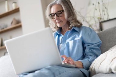 50 'li yaşlardaki gülümseyen olgun bir kadın elinde dizüstü bilgisayarla evinde oturuyor. Üst düzey mutlu iş kadını bilgisayar teknolojileri kullanıyor, çalışıyor, sohbet ediyor, sosyal medyada zaman geçiriyor, internet, ekommerce.