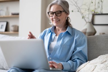 50 'li yaşlardaki gülümseyen olgun bir kadın elinde dizüstü bilgisayarla evinde oturuyor. Üst düzey mutlu iş kadını bilgisayar teknolojileri kullanıyor, çalışıyor, sohbet ediyor, sosyal medyada zaman geçiriyor, internet, ekommerce.
