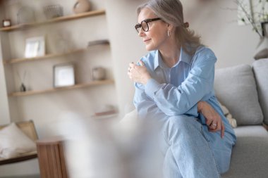 Oturma odasındaki kanepesinde dinlenen mutlu olgun bir kadın. Mavi gömlekli mutlu kadın portresi gülümsüyor..