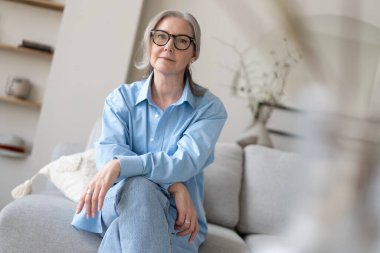 Oturma odasındaki kanepesinde dinlenen mutlu olgun bir kadın. Mavi gömlekli mutlu kadın portresi gülümsüyor..