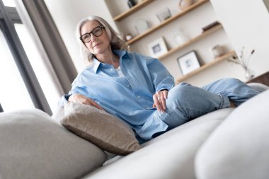 Oturma odasındaki kanepesinde dinlenen mutlu olgun bir kadın. Mavi gömlekli mutlu kadın portresi gülümsüyor..