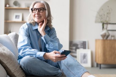 Akıllı telefondaki uygulamaları kullanarak tatlı, olgun bir bayan koltukta dinleniyor. Mutlu yaşlı kadın arkadaşlarıyla ya da çocuklarıyla kurye ya da sosyal ağda sohbet ediyor, internet mağazasından alışveriş yapıyor..