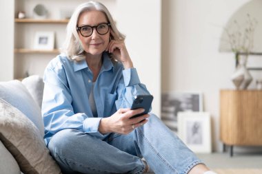 Akıllı telefondaki uygulamaları kullanarak tatlı, olgun bir bayan koltukta dinleniyor. Mutlu yaşlı kadın arkadaşlarıyla ya da çocuklarıyla kurye ya da sosyal ağda sohbet ediyor, internet mağazasından alışveriş yapıyor..