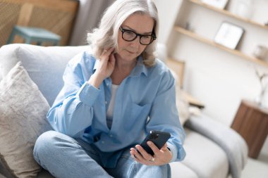 Akıllı telefondaki uygulamaları kullanarak tatlı, olgun bir bayan koltukta dinleniyor. Mutlu yaşlı kadın arkadaşlarıyla ya da çocuklarıyla kurye ya da sosyal ağda sohbet ediyor, internet mağazasından alışveriş yapıyor..