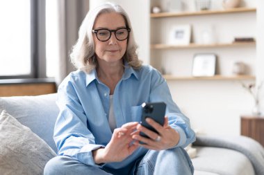 Akıllı telefondaki uygulamaları kullanarak tatlı, olgun bir bayan koltukta dinleniyor. Mutlu yaşlı kadın arkadaşlarıyla ya da çocuklarıyla kurye ya da sosyal ağda sohbet ediyor, internet mağazasından alışveriş yapıyor..