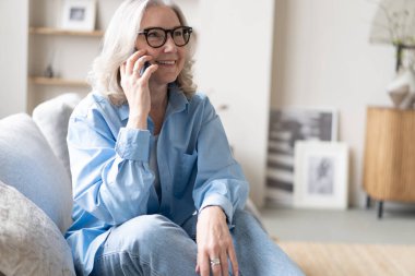 Mutlu, olgun bir kadın cep telefonu portresinden konuşuyor. Yaşlı gri saçlı bayan telefon ediyor, evdeki koltukta oturuyor, konuşmanın, iletişimin, gülümsemenin tadını çıkarıyor.